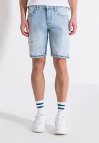 Pantalones cortos de mezclilla azul claro con detalles desgastados y dobladillo deshilachado. Llevados con zapatillas blancas y calcetines a rayas. Diseño de corte estándar.