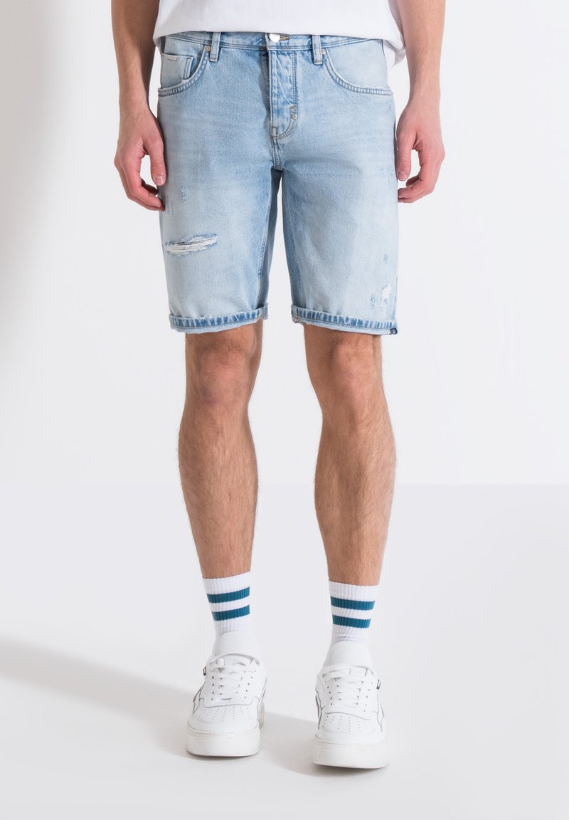 Pantalones cortos de mezclilla azul claro con detalles desgastados y dobladillo deshilachado. Llevados con zapatillas blancas y calcetines a rayas. Diseño de corte estándar.