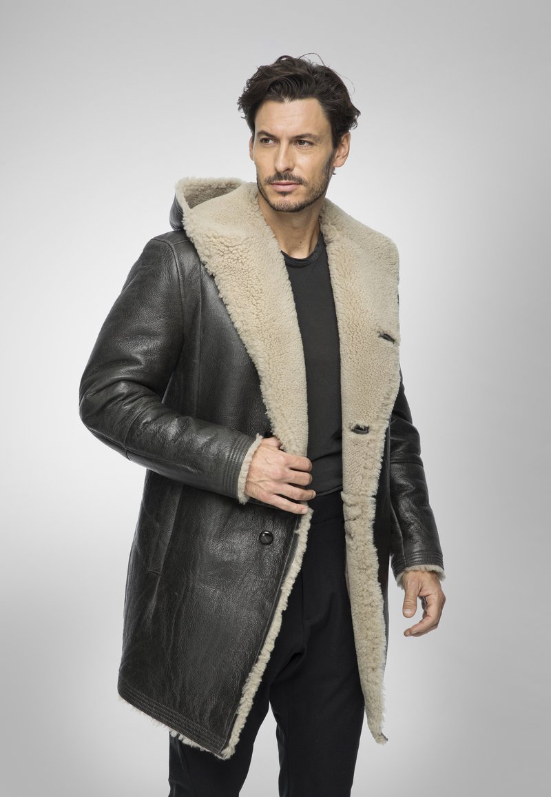 Uomo che sistema un lungo cappotto di pelle nera con fodera in shearling beige, indossando una camicia e pantaloni neri, in piedi contro uno sfondo grigio uniforme.