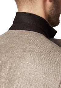 Beżowa tkana kurtka z ciemnym filcowym kołnierzem, prezentująca strukturalny fason i subtelny wzór tekstury na tkaninie.