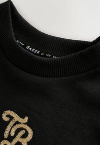 Nærbillede af en sort ribbet crewneck trøje med gylden broderet 'TB' monogram og 'Baker by Ted Baker' label indeni kraven.