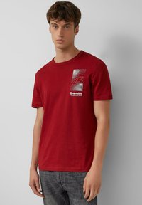 Młody mężczyzna w czerwonej koszulce z białym topograficznym wzorem oraz napisem "Terra widoczna NA NATURALNEJ ŚCIEŻCE", w połączeniu z wyblakłymi czarnymi dżinsami.