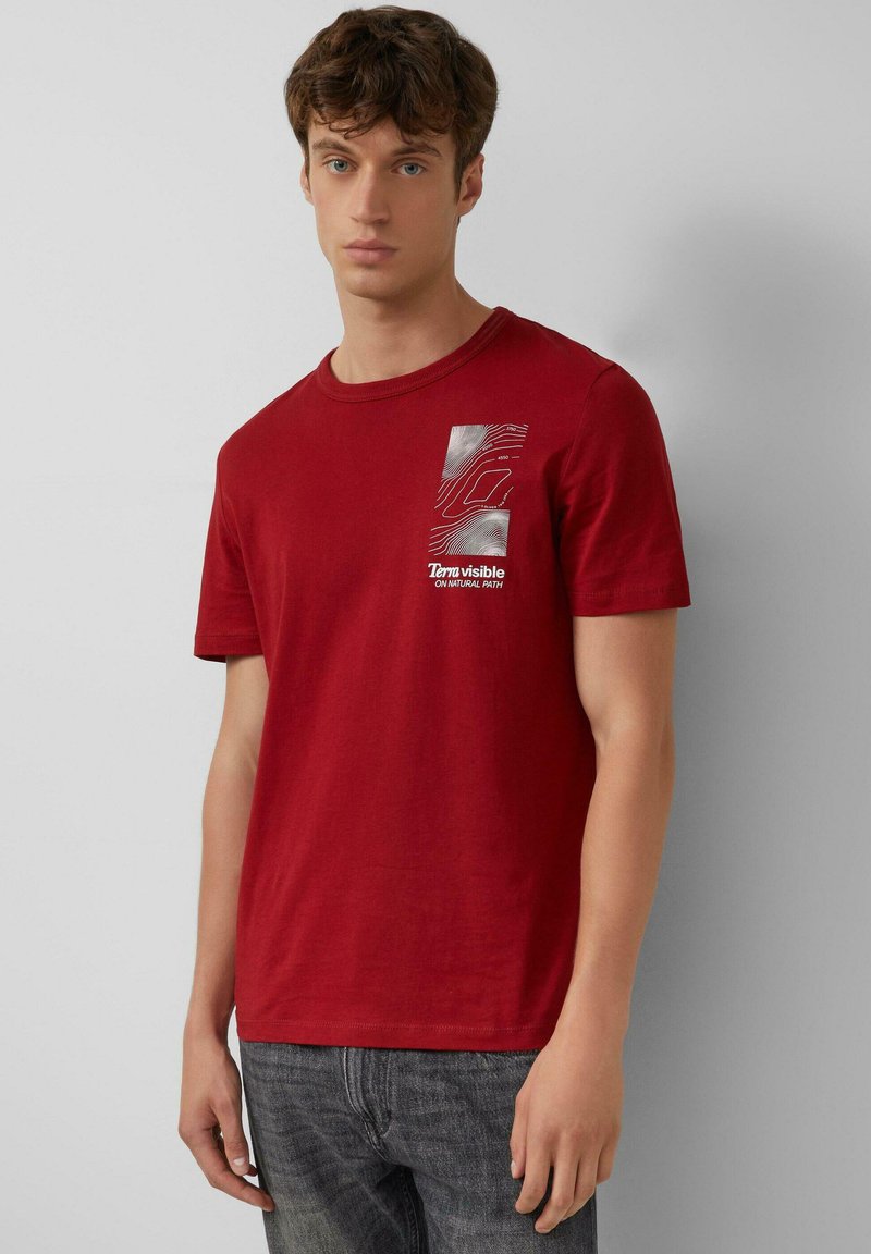 Młody mężczyzna w czerwonej koszulce z białym topograficznym wzorem oraz napisem "Terra widoczna NA NATURALNEJ ŚCIEŻCE", w połączeniu z wyblakłymi czarnymi dżinsami.