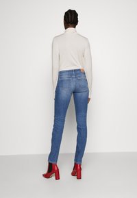 Blue denim skinny jeans with a mid-rise waist, back pockets, and a tan leather patch; paired with a beige top and red block-heeled shoes.