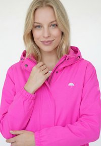 Jachetă impermeabilă roz aprins cu glugă, manșete elastice, fermoar frontal și un mic logo alb cu umbrelă pe piept.