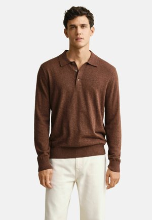 Jonge man die een bruin gebreid poloshirt met lange mouwen en een witte broek draagt, staand voor een effen witte achtergrond.