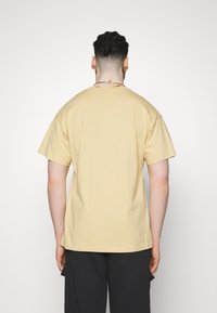 Man med kort lockigt hår iförd en beige t-shirt och svarta byxor, stående vänd mot en enfärgad vit bakgrund.