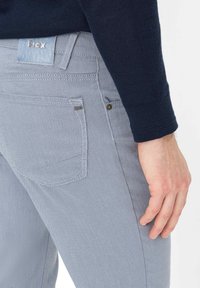 Man draagt blauwe gestructureerde broek met Brax-logo op achterzak en label, en een donkerblauwe trui, hand rustend langs het lichaam.