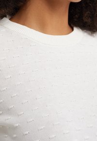 Pull en laine blanc avec un motif texturé à petits points surélevés, col rond et col côtelé. Matière douce, sans embellissements visibles.