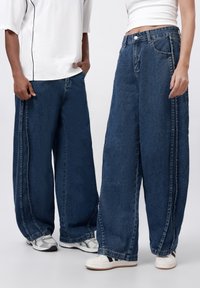 Bredvidsjeans i mörkblått, med klassisk femficksdesign och synlig sömnad längs sidorna. Bärs med avslappnade skor.