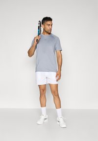 Hallutsiv hall t-särk logoga, mis on kombineeritud valgete lühikeste pükstega, millel on brändimine. Mudel hoiab tennisereketit, kandes valgeid tennisetossusid ja sokke.