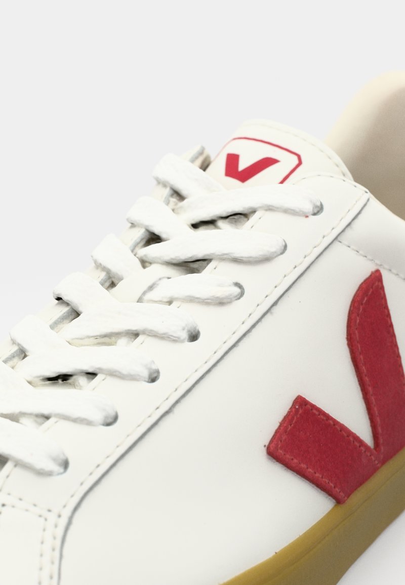 Sneaker in pelle bianca con logo N rosso, lacci bianchi e suola in gomma. Parte superiore leggermente testurizzata con una finitura liscia.