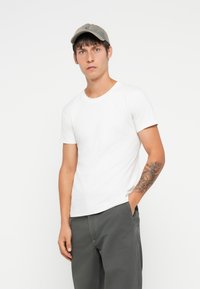 White short-sleeve t-shirt with a round neckline, paired with gray pants. The person wears a light gray cap and has a patterned arm tattoo.