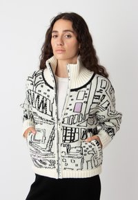 Gebreide ritscardigan met een witte basis en zwart-roze geometrische patronen, hoge kraag en twee voorzakken.