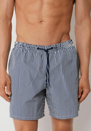Mann trägt navyblaue und weiße vertikal gestreifte Badeshorts mit Kordelzug in der Taille, steht vor einem einfarbigen Hintergrund.