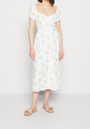 Femme portant une robe midi blanche à imprimé floral vert, manches bouffantes et encolure carrée, associée à des sandales slides tressées beiges.