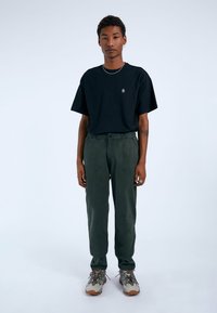 Svart t-shirt med kort ärm, lös passform, kombinerad med olivgröna tapered-byxor och grå-bruna sneakers med livliga accenter. Enkel kedjessmycke.