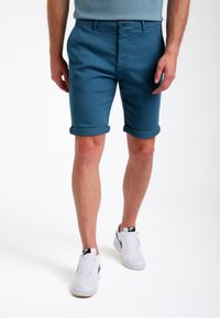 Pantaloni corti di cotone blu con orli arrotolati, tasche frontali e chiusura a bottone, abbinati a sneakers bianche con dettagli neri.
