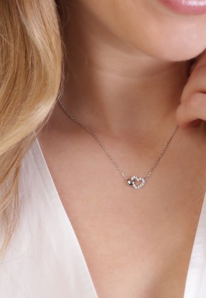 Zilveren ketting met interlocking hartvormen, de ene glad en zilverkleurig, de andere versierd met kleine heldere edelstenen.