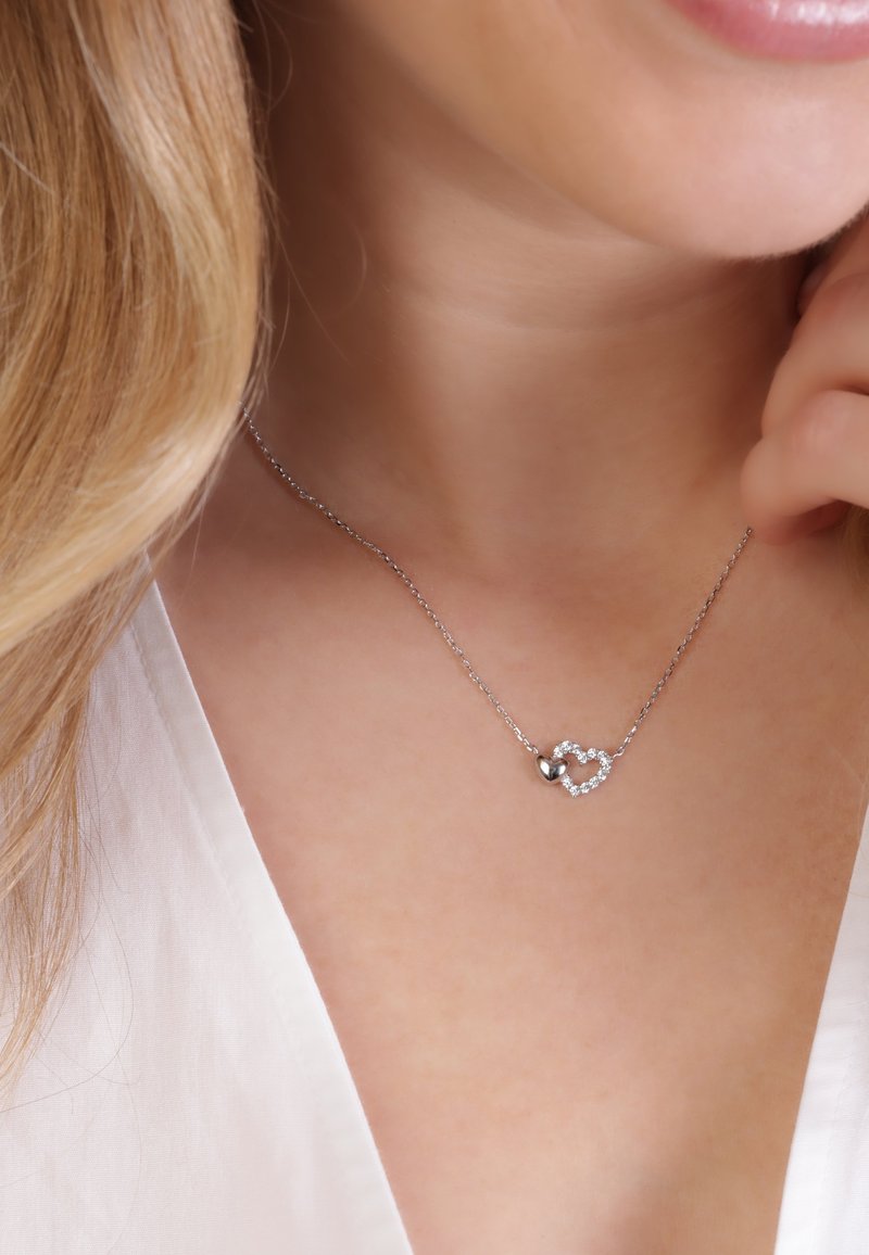 Zilveren ketting met interlocking hartvormen, de ene glad en zilverkleurig, de andere versierd met kleine heldere edelstenen.