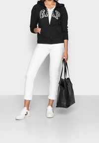 Person klädd i svart GAP-hoodie, vit skjorta, vita upprullade byxor, vita sneakers, som håller en svart läder tote-väska vid sidan.