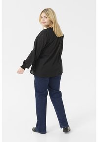 Blouse noire à manches longues avec motif texturé, encolure froncée et poignets élastiques, associée à un jean droit bleu foncé et des chaussures noires.