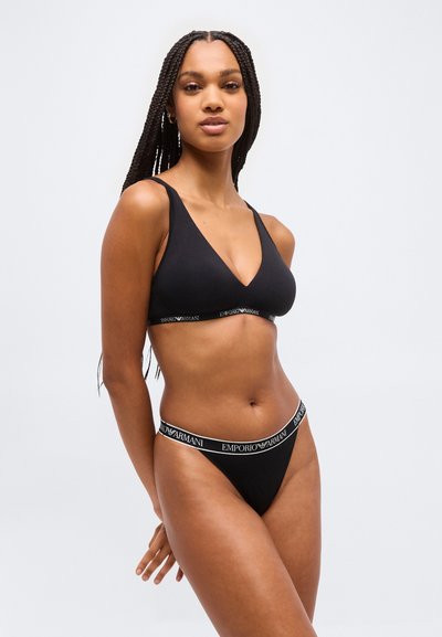 Γυναίκα με μακριά πλεξούδα που φοράει μαύρο σουτιέν με το λογότυπο της Emporio Armani και ταίριαστα σλιπ ψηλού κοψίματος, στέκεται μπροστά σε απλό φωτεινό φόντο.