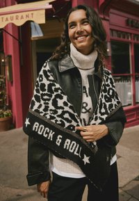Sort læderjakke over en hvid turtleneck, med et sort-hvidt leopardprint sjal der har teksten "ROCK & ROLL" og stjerneaccentuer.