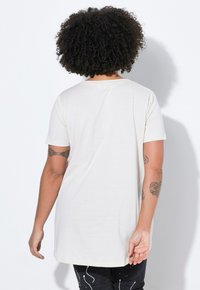 Camiseta de color crema, de manga corta y hecha de tejido suave; presenta un corte relajado y un escote redondeado, combinada con jeans oscuros estampados.