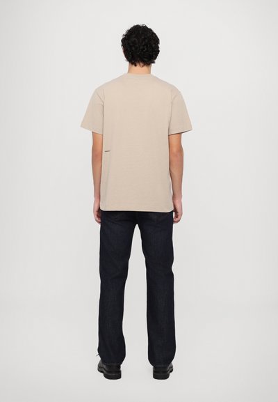 Tricou din bumbac bej, cu mâneci scurte, croială lejeră și un detaliu mic cu logo la șold, asortat cu blugi drepti de culoare albastru închis.