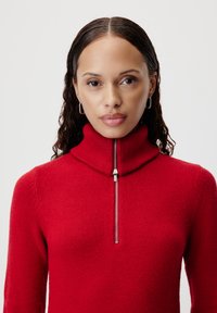 Maglione rosso a collo alto con mezza cerniera, realizzato in tessuto morbido. Presenta un colletto a costine e una texture liscia, silhouette aderente.