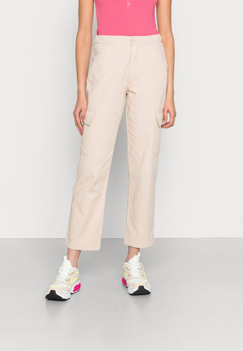 Calças cargo de um tom bege claro com dois bolsos laterais, design de perna reta e costura visível. Combinadas com ténis de desporto brancos e cor-de-rosa.