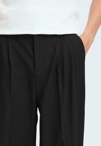 Pantalons noirs ajustés avec deux plis frontaux, poches latérales et une texture de tissu lisse, associés à un haut blanc.