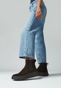 Botas de ante marrón hasta el tobillo con paneles laterales elásticos negros, suela de goma negra gruesa, combinadas con vaqueros de pierna ancha azul claro.