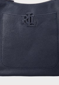 Γυναικεία τσάντα από πετρώδη δέρμα σε ναυτικό μπλε, με εμπρόσθια τσέπη και ανάγλυφο λογότυπο. Περιέχει κορυφαίο φερμουάρ και επίπεδο δομημένο σχέδιο.