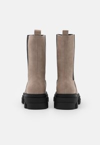 Ett par beige mockaskor med svarta klumpiga sulor, sedda bakifrån mot en vit bakgrund.