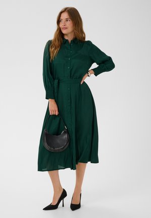 Femme portant une robe midi boutonnée à manches longues de couleur vert foncé avec une ceinture, tenant un sac à main noir et portant des talons hauts noirs.