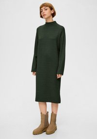 Kobieta z krótkimi włosami, ubrana w długą, zieloną sukienkę swetrową midi z długim rękawem oraz jasnobeżowe botki do kostki, stojąca na tle jednolitego tła.