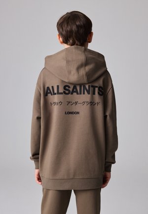 Henkilö, jolla on päällään ruskea huppari ja housut, selkä kameraan päin, hupparissa on takana painettuna "ALLSAINTS", japaninkielistä tekstiä ja "LONDON".