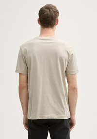 Camiseta de manga corta beige claro con cuello redondo, con una textura de tela suave y un corte recto, mostrada desde la parte de atrás.
