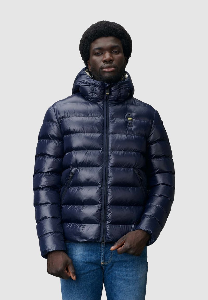 Giacca imbottita blu navy con cappuccio, dotata di zip frontale, due tasche laterali e una texture liscia e lucida. Vestibilità standard con design a quadri orizzontali.