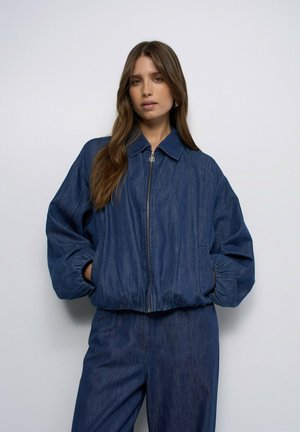 Mujer con una chaqueta vaquera azul oscuro holgada con cremallera y puños elásticos, y pantalones vaqueros de cintura alta a juego sobre un fondo liso.