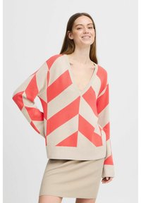 Pull en chevrons corail et beige avec un col en V profond, coupe loose et manches longues. Ourlet inférieur côtelé, associé à une jupe en tricot beige.
