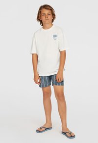 Witte T-shirt met een klein blauw logo, gecombineerd met marineblauwe shorts met patroon en blauwe flip-flops. Casual, ontspannen fit en comfortabele materialen.