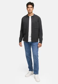 Mörkgrå skjorta med knappar och två bröstfickor, bärs över en vit t-shirt, kombinerad med blå jeans och vita sneakers.