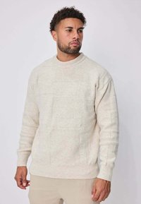 Pull léger beige avec un motif texturé. Il a un col rond, des poignets et un ourlet côtelés. Matière douce en maille avec une coupe décontractée.