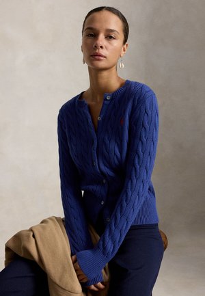 Frau mit zurückgebundenen Haaren, die eine blaue, kabelgestrickte Strickjacke und marineblaue Hose trägt, hält einen beigen Mantel und sitzt vor einem schlichten neutralen Hintergrund.