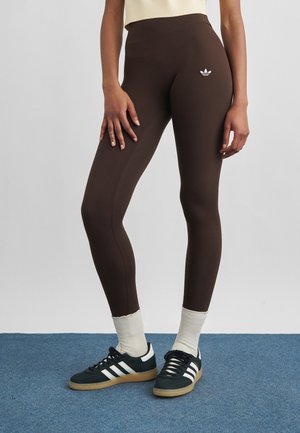 Femme portant un legging Adidas marron foncé avec un logo sur la cuisse, des chaussettes blanches et des baskets Adidas noires avec des bandes blanches sur un tapis bleu.