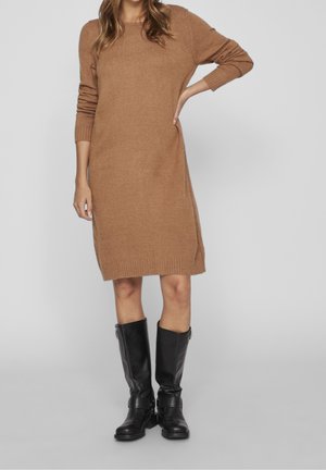 Vrouw die een camelkleurige, knielange trui-jurk draagt met lange mouwen en zwarte leren laarzen tot aan de knie, staande met één hand op haar heup.