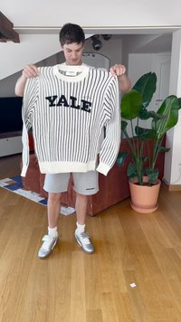 Baltas megztinis su juodais vertikaliais dryžiais ir storais "YALE" užrašais. Dėvimas ant pilkų šortų; modelis laiko megztinį ryškiai apšviestoje patalpoje.